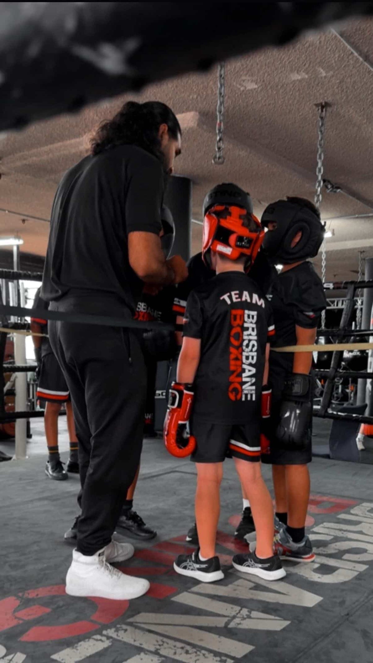 A Brisbane Boxing coach giving instructions to a group of young boxers in headgear and Team Brisbane Boxing kit beside the ring.